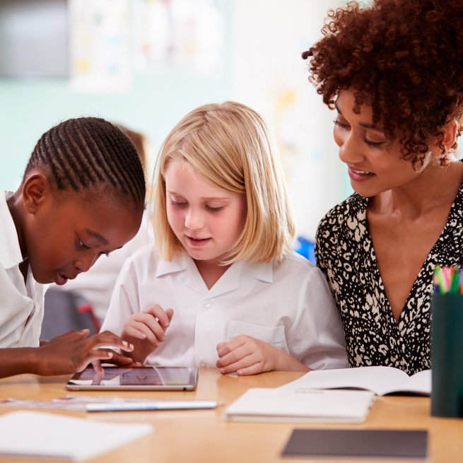 A teacher looks on as her two students interact with a tablet.